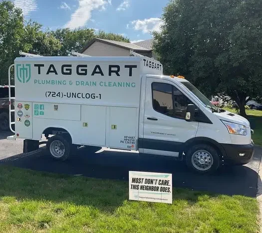 Side view of a white Taggart Plumbing service van with branded yard sign at a residential home in Monaca, PA